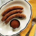 A plate with three grilled sausages and a small dish of sauce on the side, next to a yellow napkin and a fork.