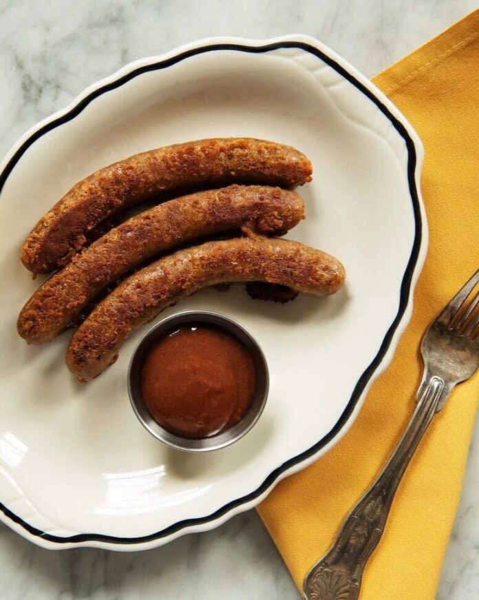 Sticky Little Sausages A plate with three grilled sausages and a small dish of sauce on the side, next to a yellow napkin and a fork.