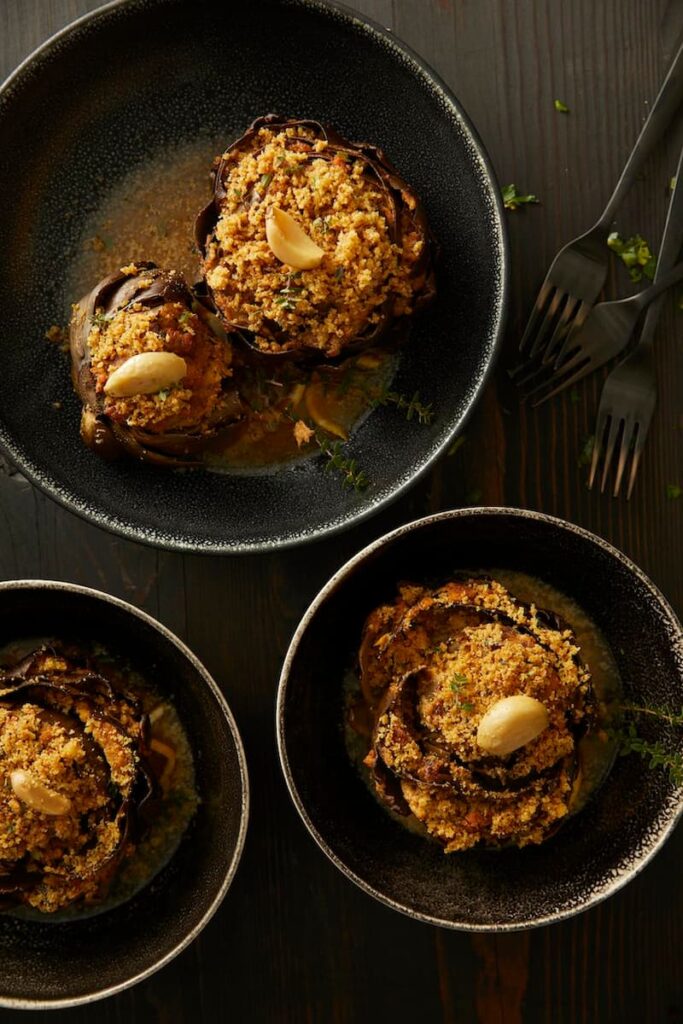 Three stuffed artichokes in black bowls, with a garlic clove garnish and a sauce in the bottom of the bowl.