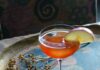 The Ichabod Crane Cocktail A reddish-orange cocktail in a coupe glass, garnished with a thin apple slice, and resting on a rustic serving tray with dried foliage.