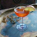 A reddish-orange cocktail in a coupe glass, garnished with a thin apple slice, and resting on a rustic serving tray with dried foliage.