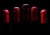 The Sexiest of Grapes: Syrah A row of five dark red wine bottles with red foil caps, positioned closely together against a black background.