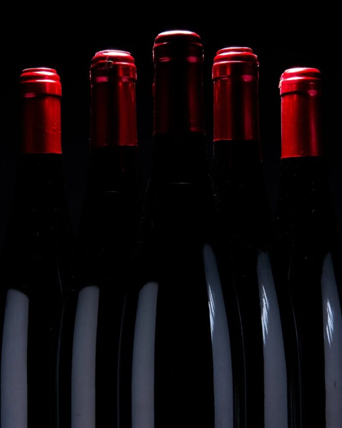 A row of five dark red wine bottles with red foil caps, positioned closely together against a black background.