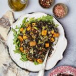 A plate of wild rice with butternut squash, raisins, and hazelnuts, served over a bed of arugula, next to a bottle of olive oil and bowls of salt and hazelnuts.