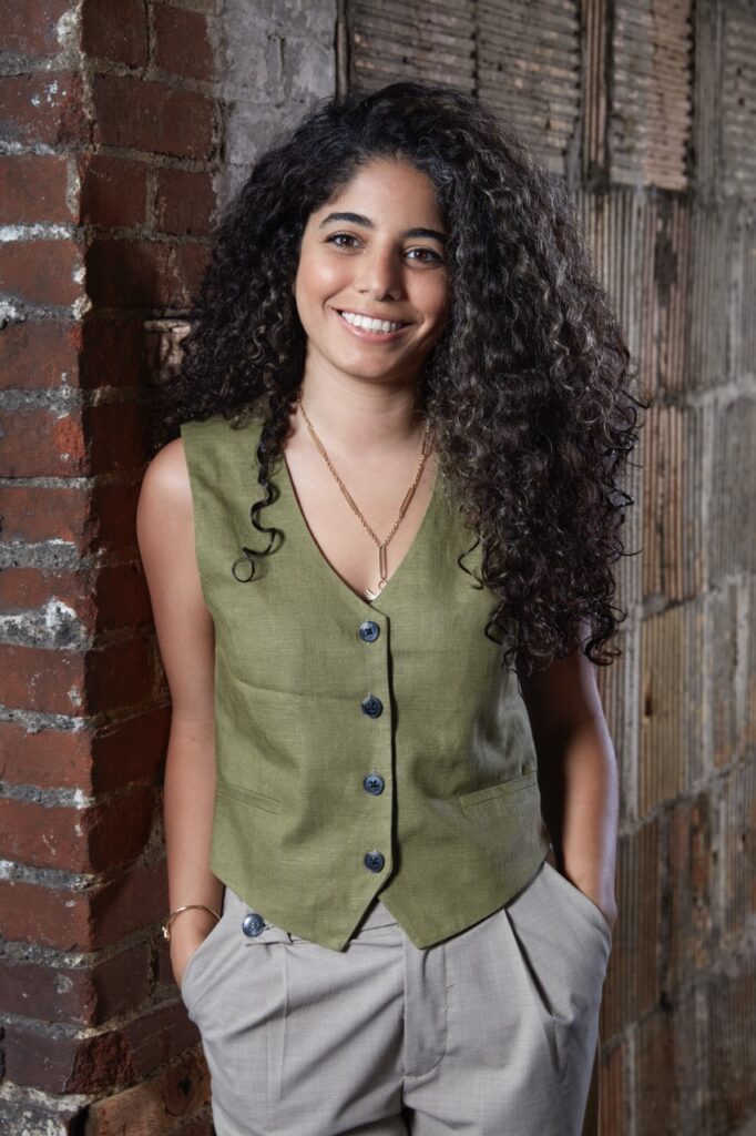 Yasmine El Meleegy in green vest smiling by brick wall