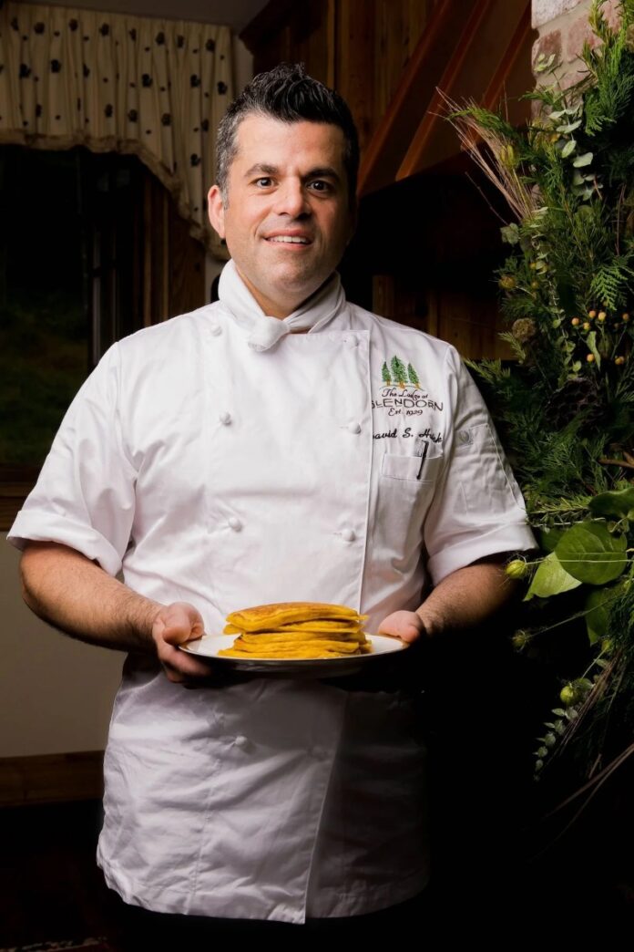 Chef David Haick in a white uniform holds a plate of golden-brown pancakes.