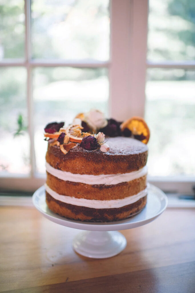 A rustic layered cake on a white cake stand, topped with dried orange slices and dark berries, sits in front of a bright window.