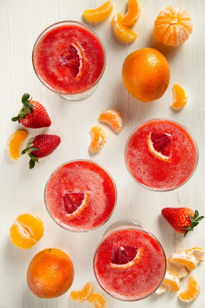 Glasses of a red cocktail garnished with a blood orange slice, surrounded by strawberries, oranges, and mandarin orange slices on a white surface.