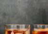 Ancho Chile Old Fashioned A moody shot of two glasses of Ancho Chile Old Fashioned cocktails on a bar, with scattered sugar crystals on the surface.