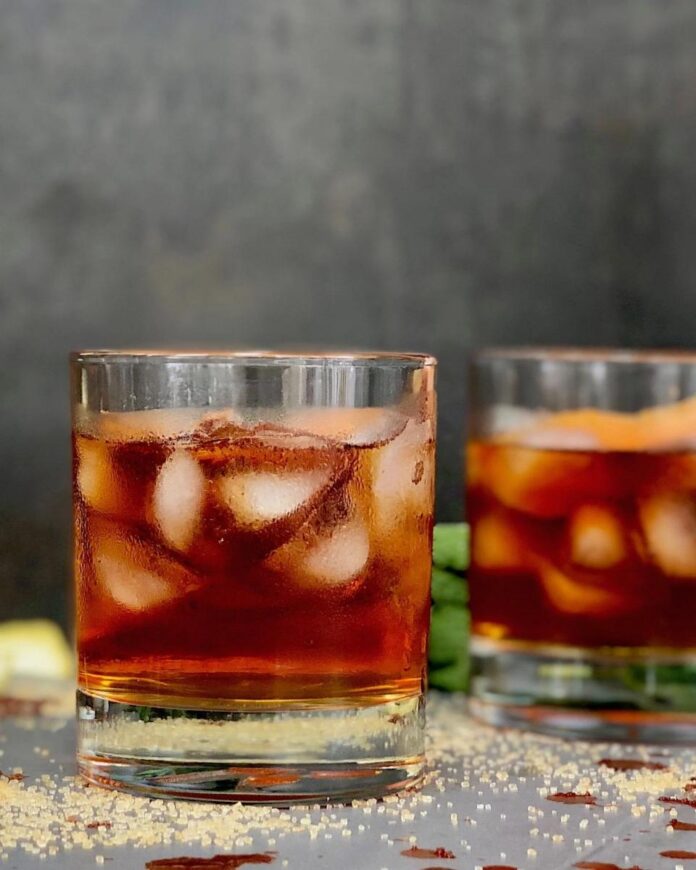 A moody shot of two glasses of Ancho Chile Old Fashioned cocktails on a bar, with scattered sugar crystals on the surface.
