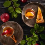A warm Apple Cinnamon Hot Toddy in a clear mug, garnished with an apple slice and a cinnamon stick, served with a slice of cake on a brown plate.