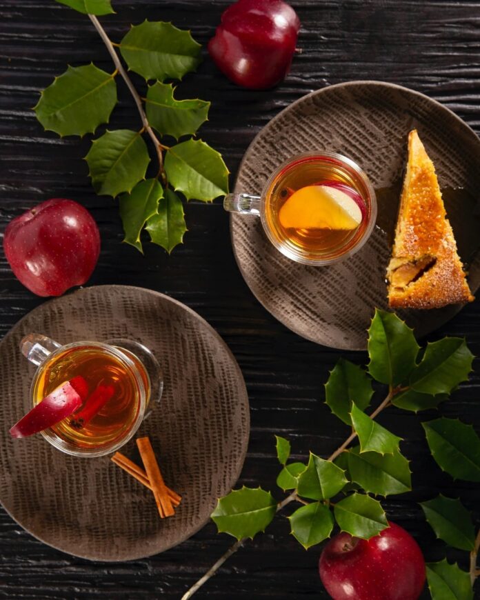 Apple Cinnamon Hot Toddy A warm Apple Cinnamon Hot Toddy in a clear mug, garnished with an apple slice and a cinnamon stick, served with a slice of cake on a brown plate.