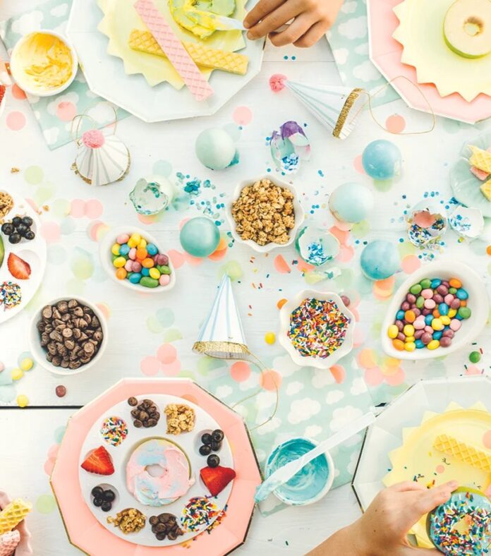 Apple Donuts TABLE Magazine A colorful overhead view of a table with plates of sliced apples decorated to look like donuts with various toppings, with bowls of candy and sprinkles, and children's hands reaching for the food.