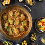 A Spanish paella dish with chicken, rice, and herbs, served in a large pan on a dark stone surface, surrounded by small bowls of various tapas and garnishes.