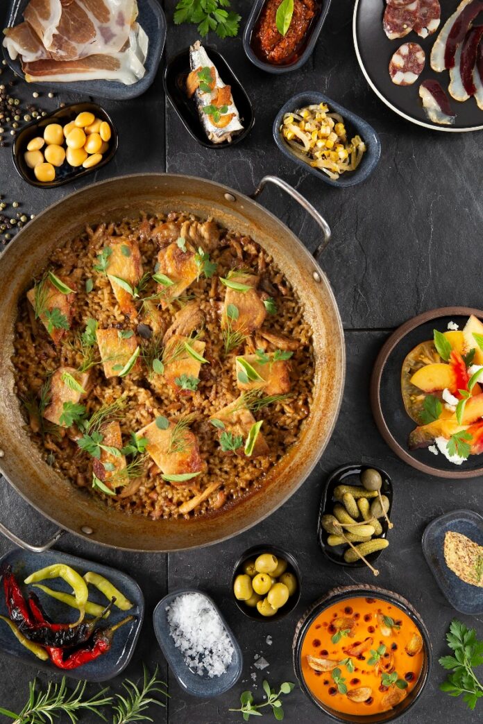 A Spanish paella dish with chicken, rice, and herbs, served in a large pan on a dark stone surface, surrounded by small bowls of various tapas and garnishes.