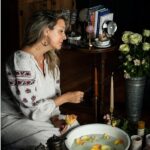 A woman in a white dress sits on a wooden floor, lighting a candle next to a large basin of water with rose petals floating in it.