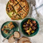 A table set with a delicious breakfast casserole, complemented by bread, figs, and an assortment of fresh fruits for sharing.