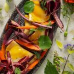 A plate of carrot pierogies garnished with carrot ribbons and fresh herbs, surrounded by flowers and green and red vegetables.