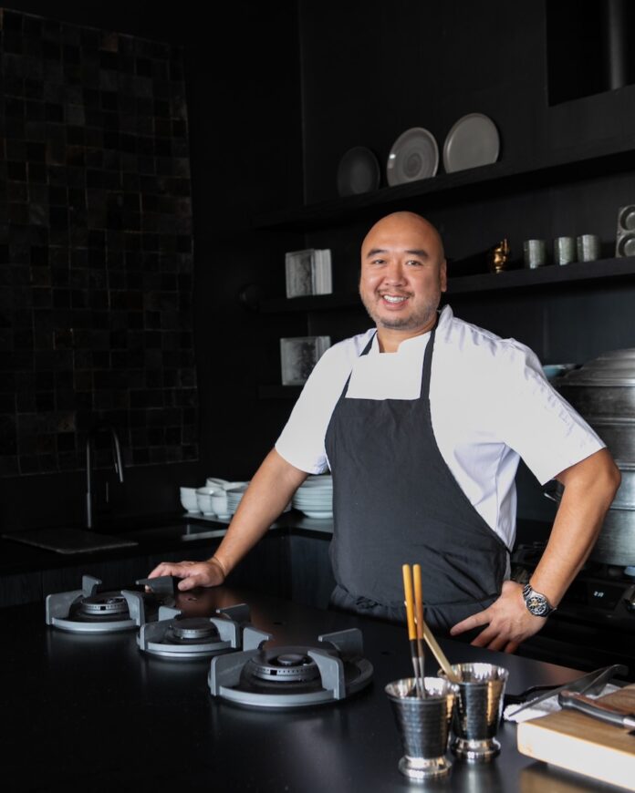 Chef Roger Li stands at a kitchen counter and stove top with an apron on, ready to make dinner.