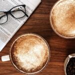 Two mugs of cinnamon spice lattes on a wooden table next to a newspaper and a small bowl of coffee beans.