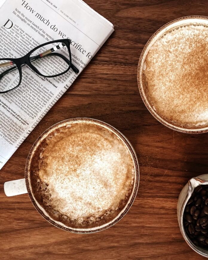 Cinnamon Spice Latte Two mugs of cinnamon spice lattes on a wooden table next to a newspaper and a small bowl of coffee beans.