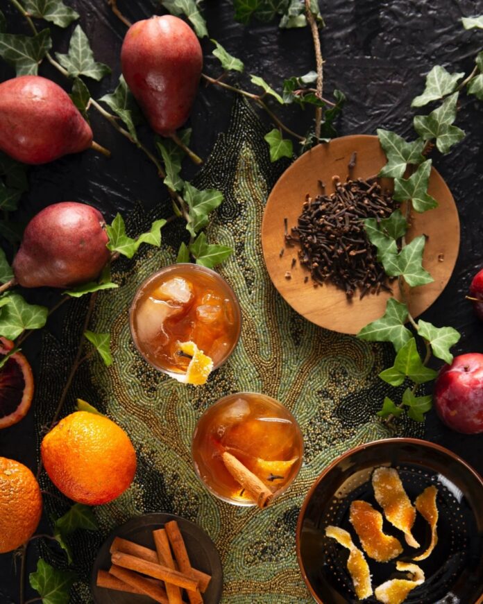 Clove Old Fashioned A Clove Old Fashioned cocktail in a rocks glass, garnished with a candied orange peel and a cinnamon stick, with cloves, cinnamon, and oranges on a beaded placemat.