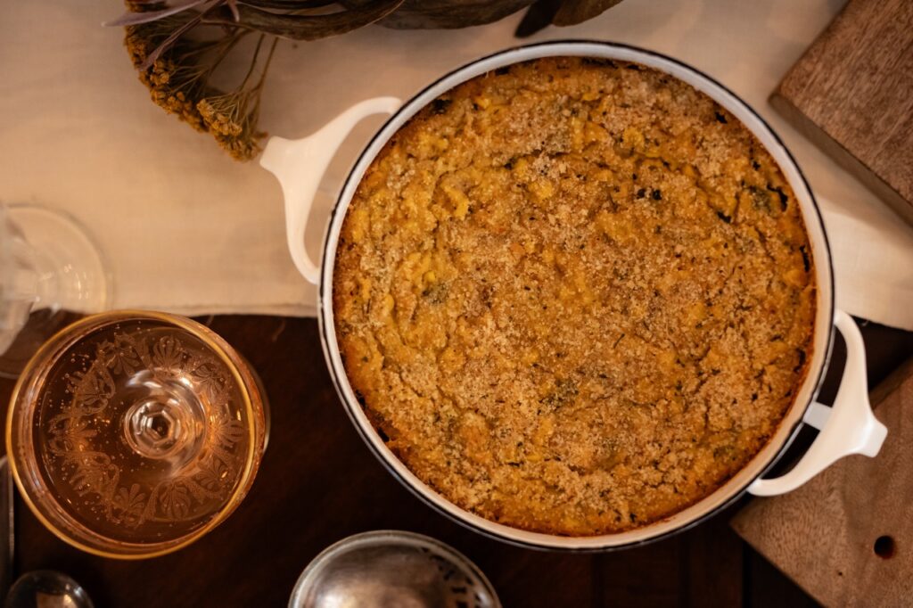 A round pot of corn casserole on a table next to a drink.