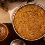 A round pot of corn casserole on a table next to a drink.