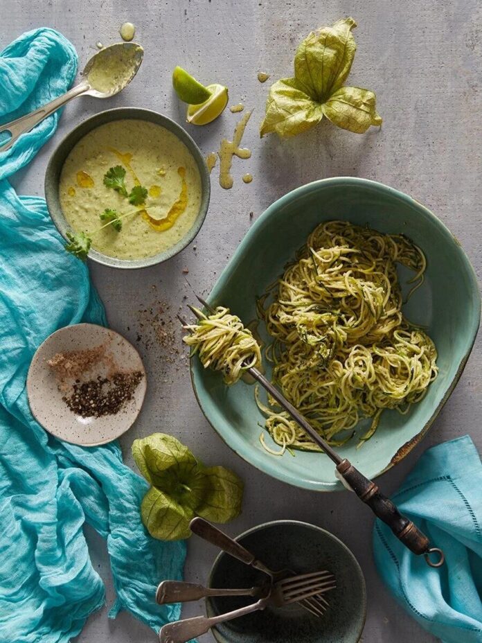 A bowl of zucchini noodles on a blue plate, with a fork holding some noodles above the bowl, alongside a bowl of zucchini soup and other ingredients on a gray table.