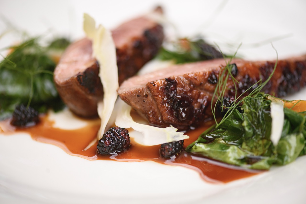 Two duck breasts sit on a white plate with fennel and a blackberry sauce.