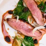 Slices of duck breast sit on a white plate surrounded by greens and a blackberry sauce.