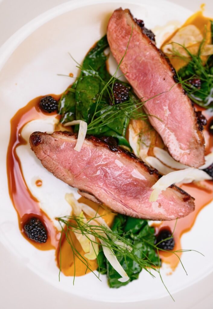 Slices of duck breast sit on a white plate surrounded by greens and a blackberry sauce.