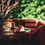 A glass-topped outdoor table with two slices of red devil's food cake on green plates, with a cup and saucer, and a plant in a white pot, against a soft green background of bushes.