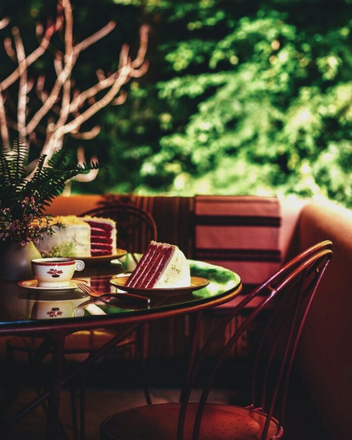 A glass-topped outdoor table with two slices of red devil's food cake on green plates, with a cup and saucer, and a plant in a white pot, against a soft green background of bushes.