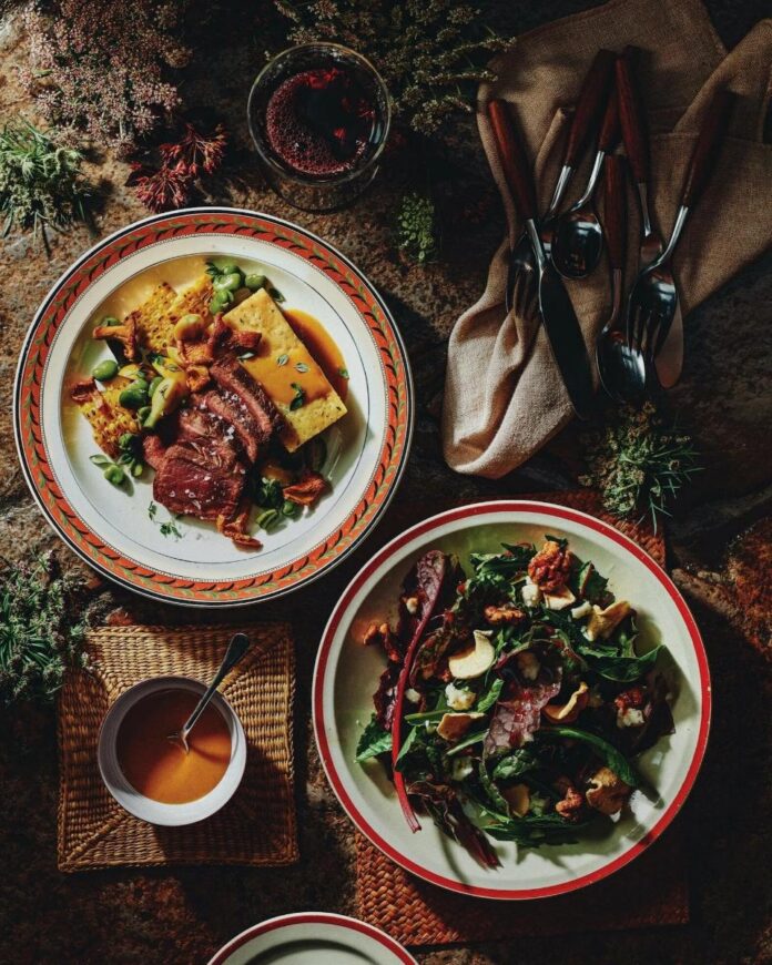 A table with two place settings and a small bowl of sauce. Each plate holds a meal with roasted venison, wild garlic polenta, vegetables, and sauce.