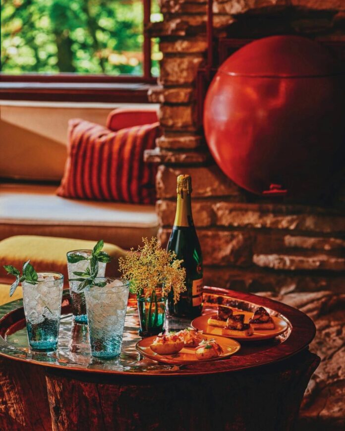 A tray with three glasses of a sparkling cocktail with mint sprigs and ice, with a bottle of champagne and plates of appetizers on a wooden table, in a cozy, sunlit room.