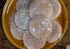 New Mexico Biscochitos A brown platter of round, scalloped-edged biscochito cookies dusted with powdered sugar, with some sugar sprinkled on the wooden table beside the platter.