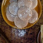 A brown platter of round, scalloped-edged biscochito cookies dusted with powdered sugar, with some sugar sprinkled on the wooden table beside the platter.