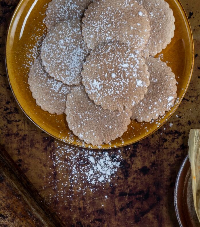 A brown platter of round, scalloped-edged biscochito cookies dusted with powdered sugar, with some sugar sprinkled on the wooden table beside the platter.