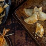 Several golden brown empanadas on a rustic metal baking sheet, with one broken open to show the filling.