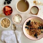 A breakfast bowl with baked carrot cake oatmeal, surrounded by small bowls of toppings, including strawberries, blueberries, blackberries, shredded coconut, and brown sugar.
