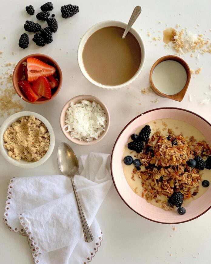 Jen Clark's Carrot Cake Baked Oatmeal TABLE Magazine A breakfast bowl with baked carrot cake oatmeal, surrounded by small bowls of toppings, including strawberries, blueberries, blackberries, shredded coconut, and brown sugar.
