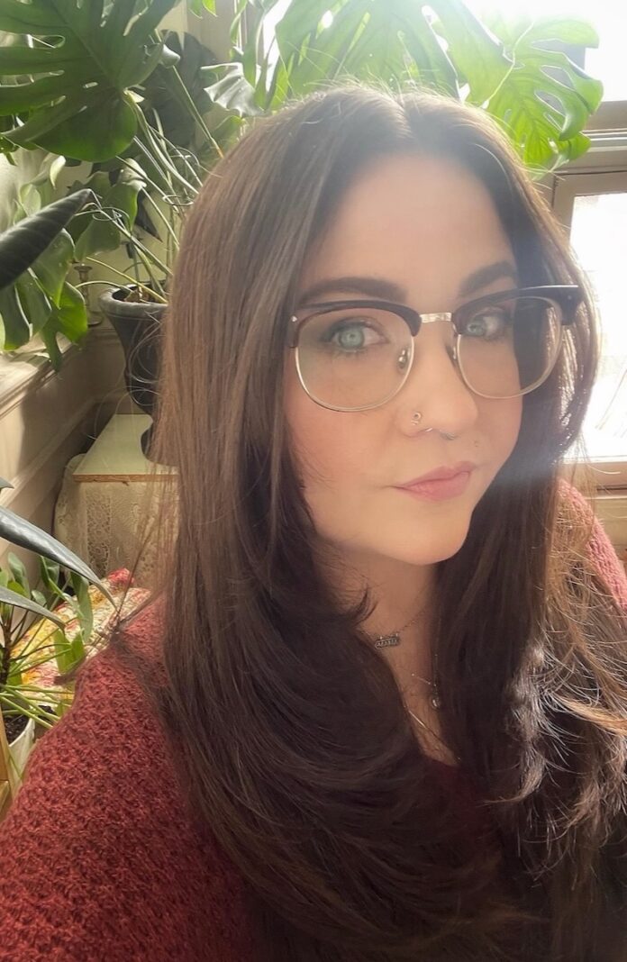 A woman in a red shirt and glasses stands in front of plants.