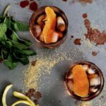 Two cocktails in rocks glasses garnished with large orange peels, next to sprigs of fresh mint and a messy pile of sugar and liquid splatters.