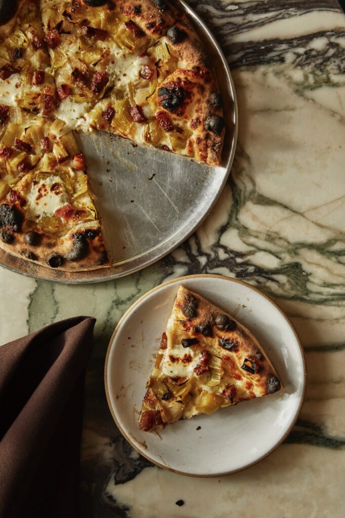 A slice of pizza taken out of a whole pizza sits on a plate below the whole pizza.