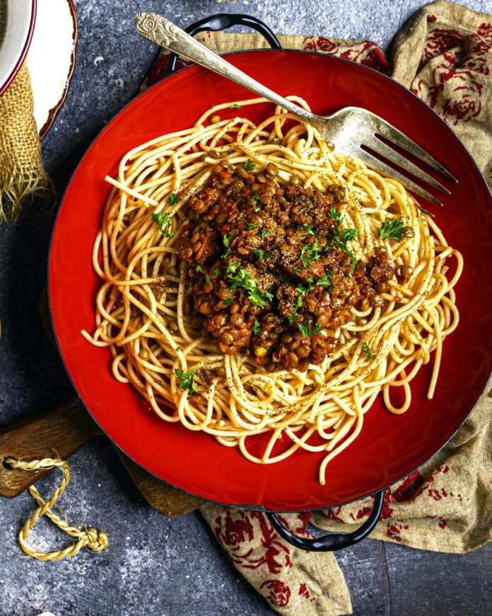 A red bowl filled with spaghetti topped with lentil bolognese sauce and parsley, with a fork resting on the side.