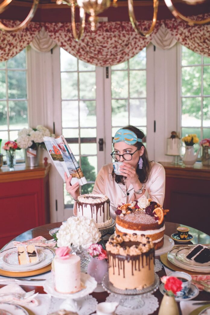 A girl wearing an eye mask and pajamas sips from a teacup while reading a magazine at a table laden with several decorated layer cakes and small plates of cake slices.