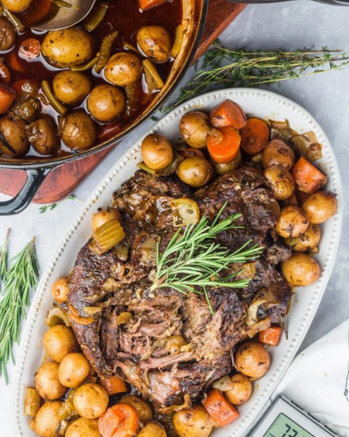A slow-cooked beef brisket on a white platter surrounded by roasted potatoes and carrots.