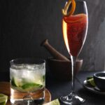 A glass of Negroni Sbagliato garnished with an orange twist next to a Caipirinha cocktail in a rocks glass with a wooden muddler and a small bowl of sugar.