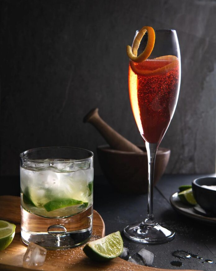 A glass of Negroni Sbagliato garnished with an orange twist next to a Caipirinha cocktail in a rocks glass with a wooden muddler and a small bowl of sugar.